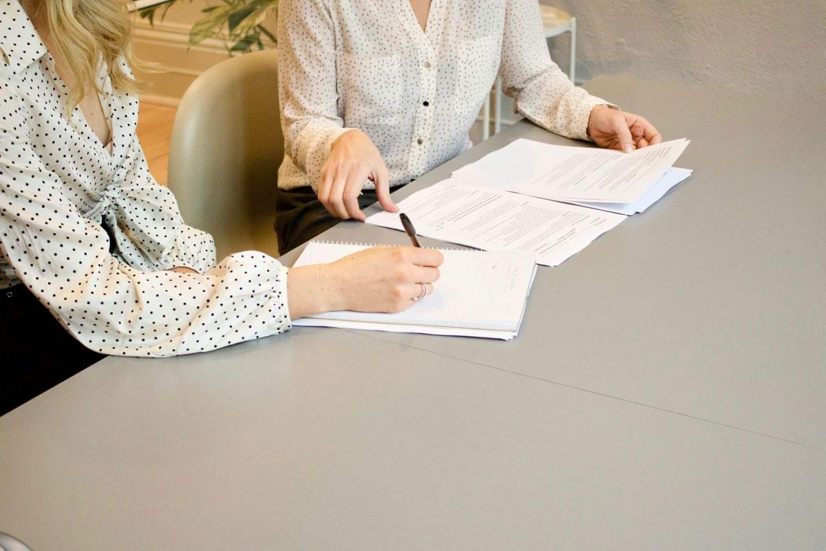 woman signing on white printer paper beside woman about to touch the documents