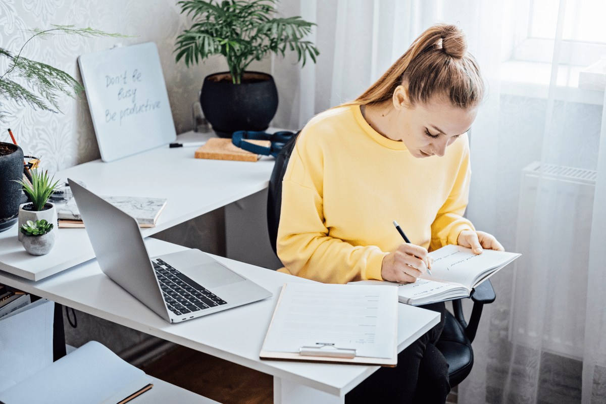 A woman in a light yellow sweater sitting at a work desk with a laptop, writing in a notebook