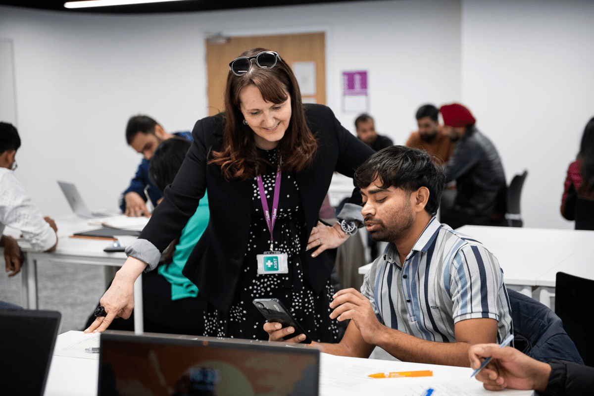 Instructor leans over a student at a desk, guiding him as he looks at his phone while others work nearby.