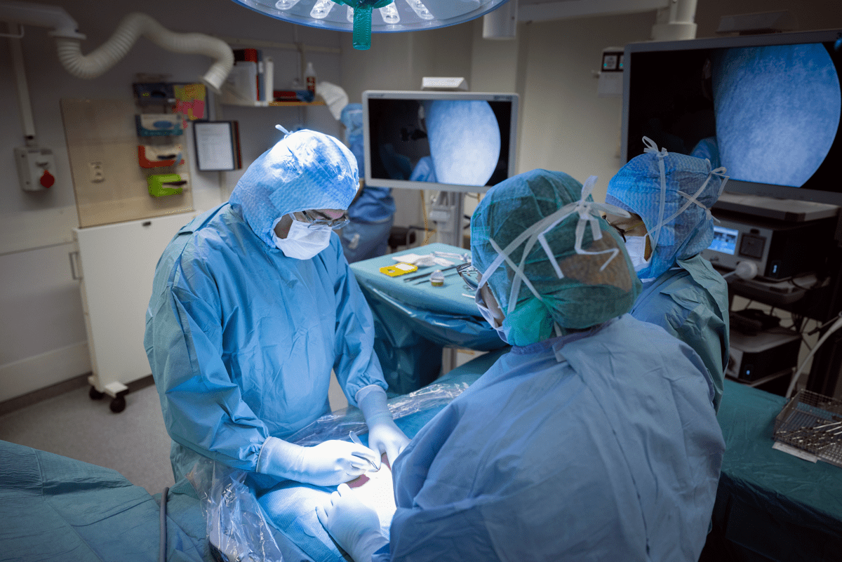 Operationssal med tre sjukvårdspersonal i skyddskläder som genomför en kirurgisk operation under stark operationsbelysning, med medicinska monitorer och utrustning i bakgrunden.