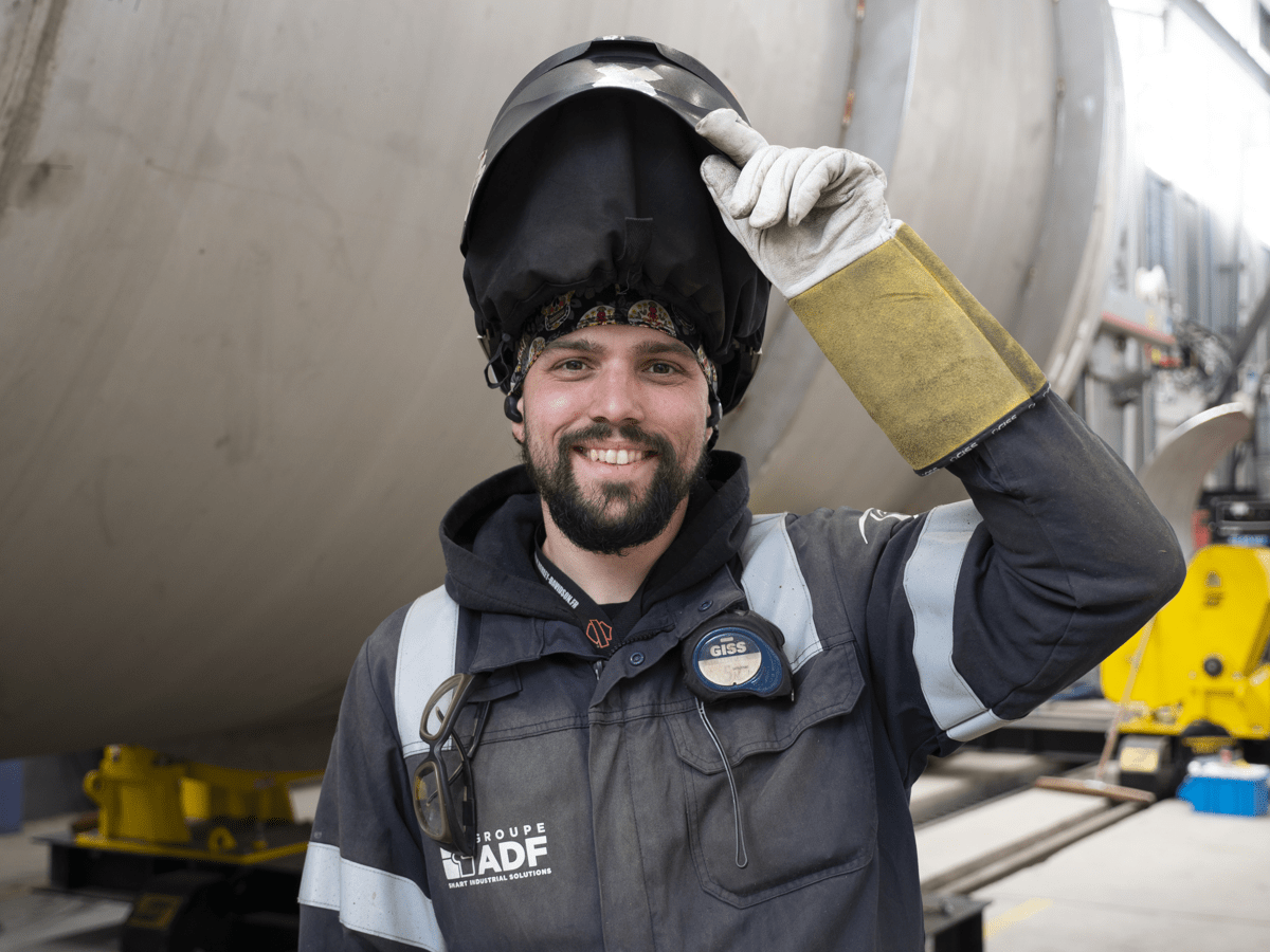 Lachend portret van een medewerker van Groupe ADF in een industriële werkplaats, met een omhooggeklapte lashelm en beschermende kleding, voor een grote metalen constructie.