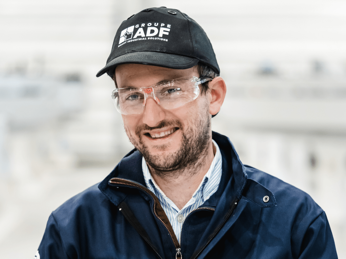 Retrato sonriente de un empleado del Groupe ADF con ropa de trabajo, una gorra negra, gafas de seguridad y una chaqueta azul marino.