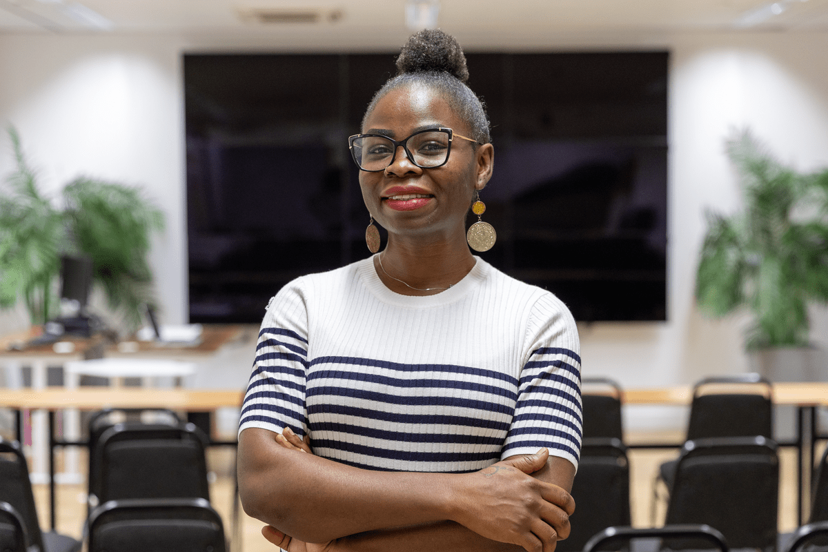 Woman in glasses and striped top standing with arms crossed in a classroom-style space.