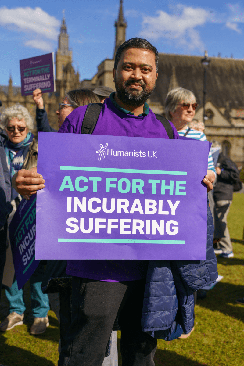 A person stands in front of the Houses of Parliament holding a Humanists UK banner saying 'Act for the incurably suffering', at the Assisted Dying Rally in May 2024