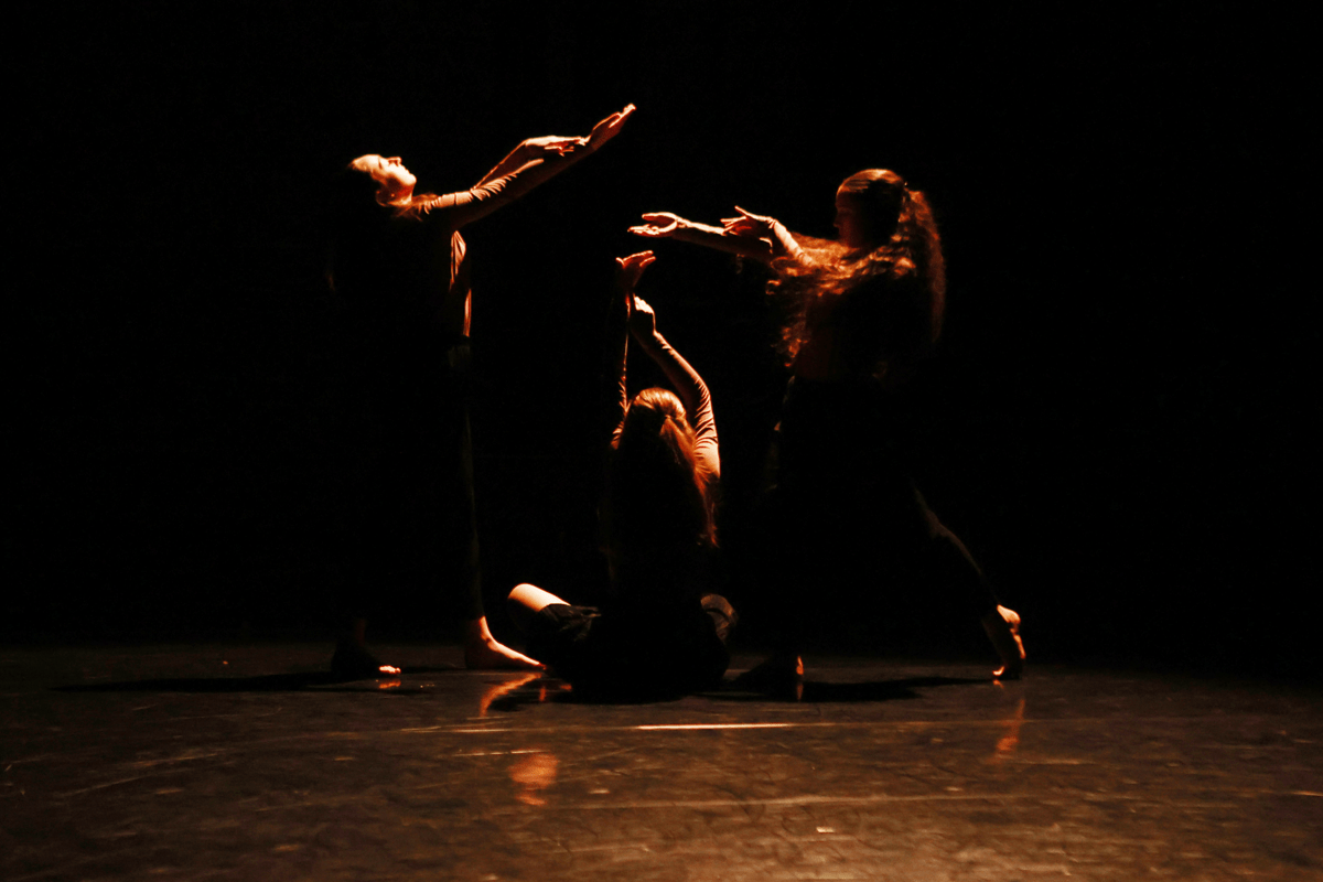 Three dancers in a blackbox stage, dimly lit. Each dancer is reaching up to the sky with palms open in a begging pose.