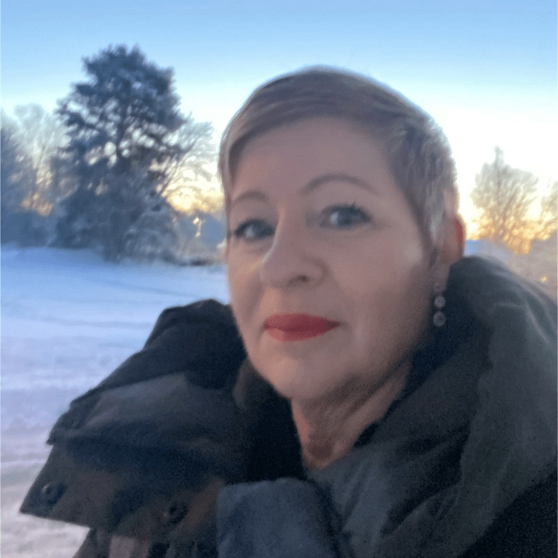 Kvinna med kort, ljusbrunt hår och röd läppstift, klädd i mörk vinterjacka. Hon står utomhus i ett vinterlandskap med snö, träd och soluppgång i bakgrunden.