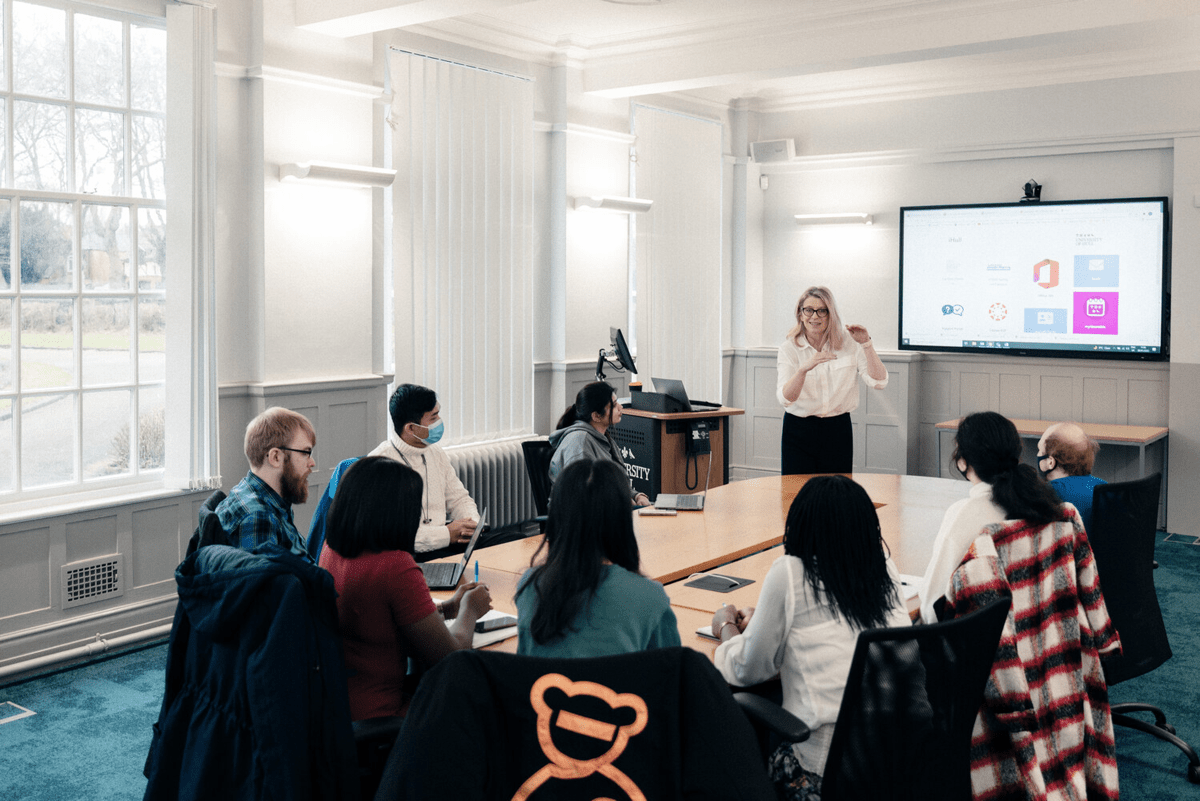 A presenter gestures while addressing a group of seated individuals around a large oval table in a meeting room. A large screen displays a presentation with various icons.