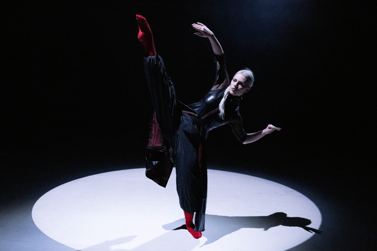 Lone student dancing on stage under a spotlight, striking a pose with one leg and arm in the air, balance on the other leg.