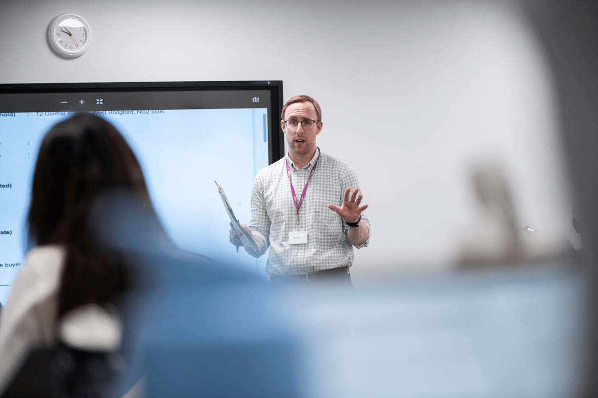 The image features a man standing in front of a large screen, possibly a TV or a projector screen, giving a presentation or lecture.