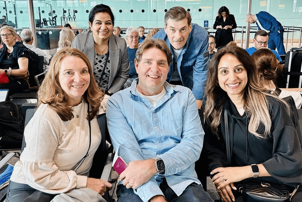A mixed group of TMF colleagues seated in an airport lounge smiling at the camera.