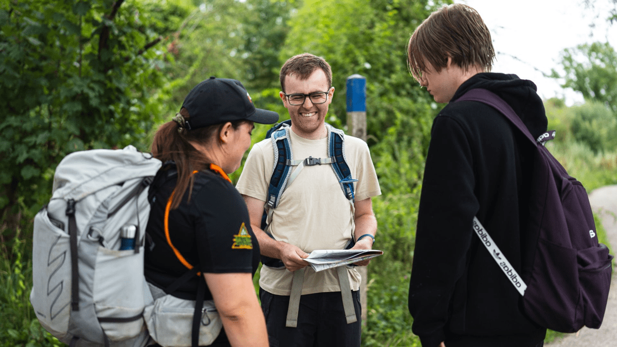 Three people wearing backpacks stand on a countryside path looking at a map together, surrounded by green trees and vegetation.