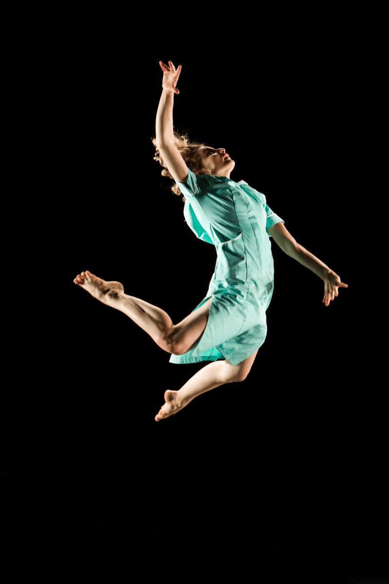 A dancer against a black background leaping in the air with splayed limbs, with long fair hair, wearing a loose fitting, turquoise smock dress.