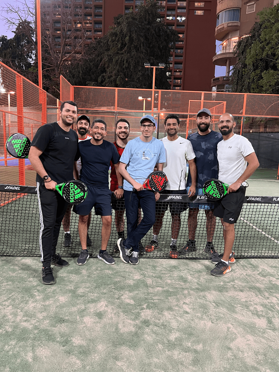 Group of friends posing on a padel court at dusk, holding padel rackets in front of the net with city buildings behind.