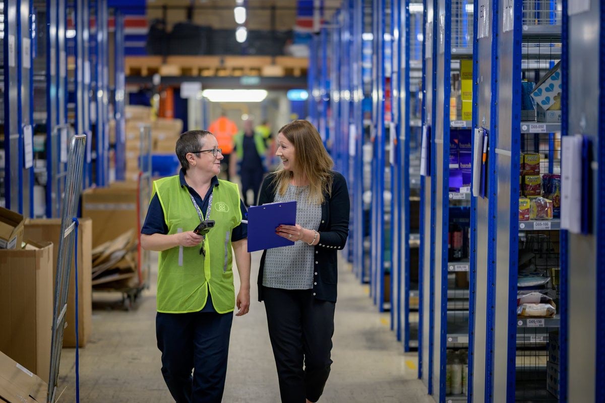 Two women walking through a warehouse and talking. One is holding a clipboard.