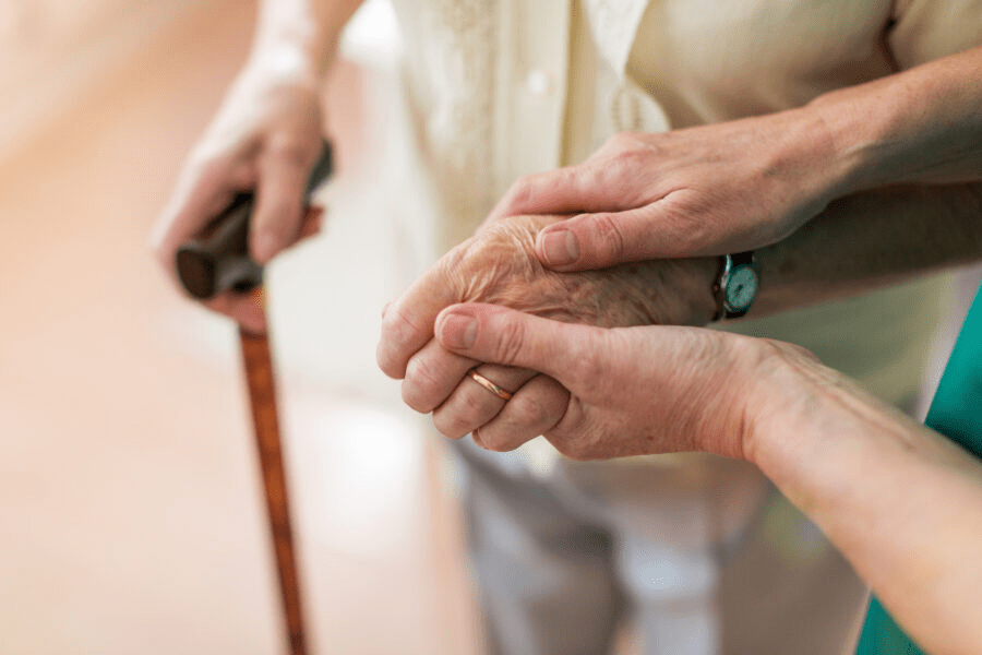 The body and legs of an older woman with a walking stick. Here hand is being help by younger looking hands