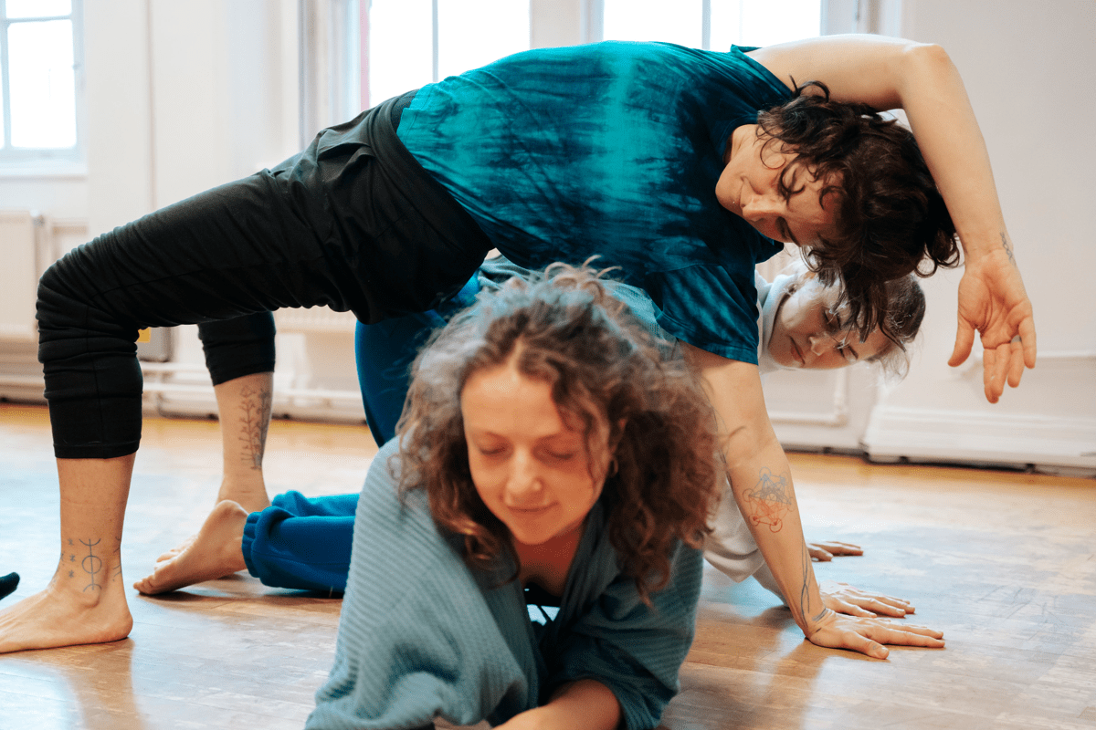 Dancers in a studio, with one dancer on the floor on their stomach, and two dancers stretched over them