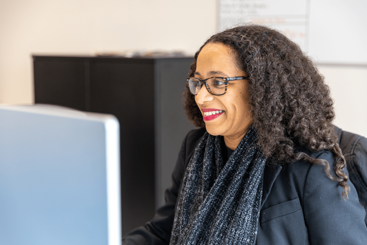 Woman with curly hair and glasses smiling while working at a computer.