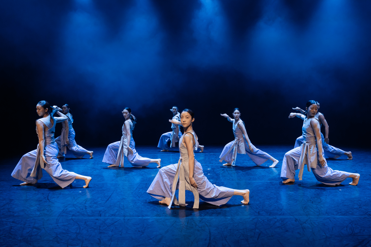 Eight dancers spread across the stage in a half-seated position with blue atmospheric lights.