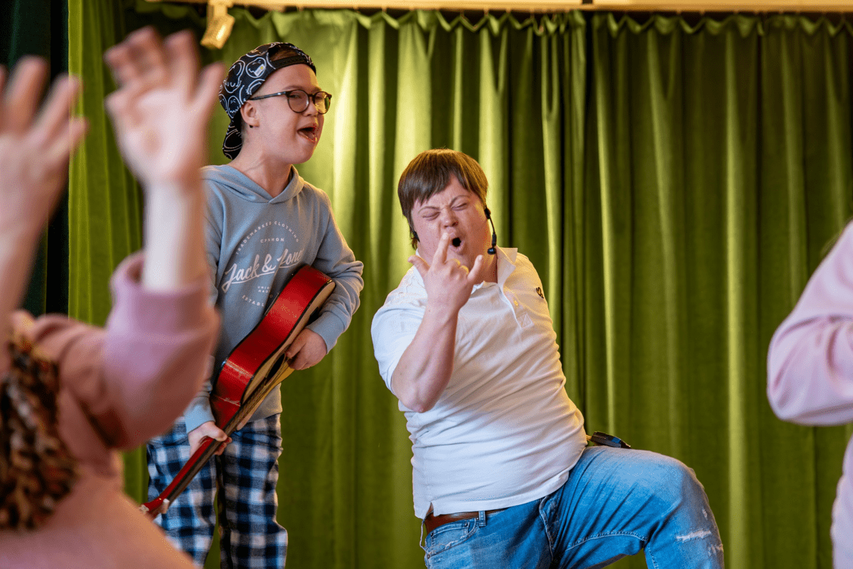 	Två unga personer, en med gitarr, sjunger och gör rockposer framför gröna gardiner.