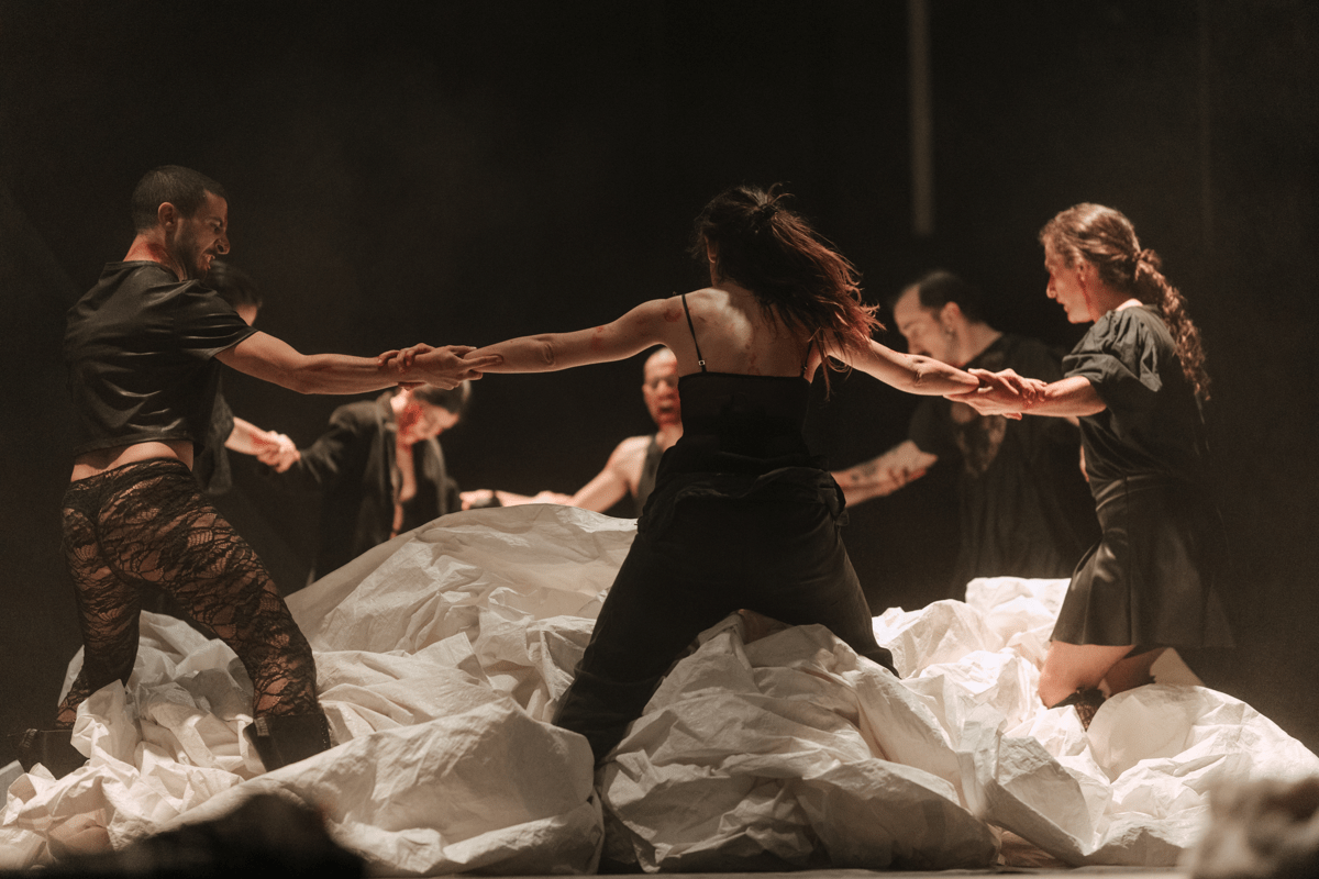 Image of a group of dancers dressed in black, in a circle holding hands, standing amongst ruffled white cloth.