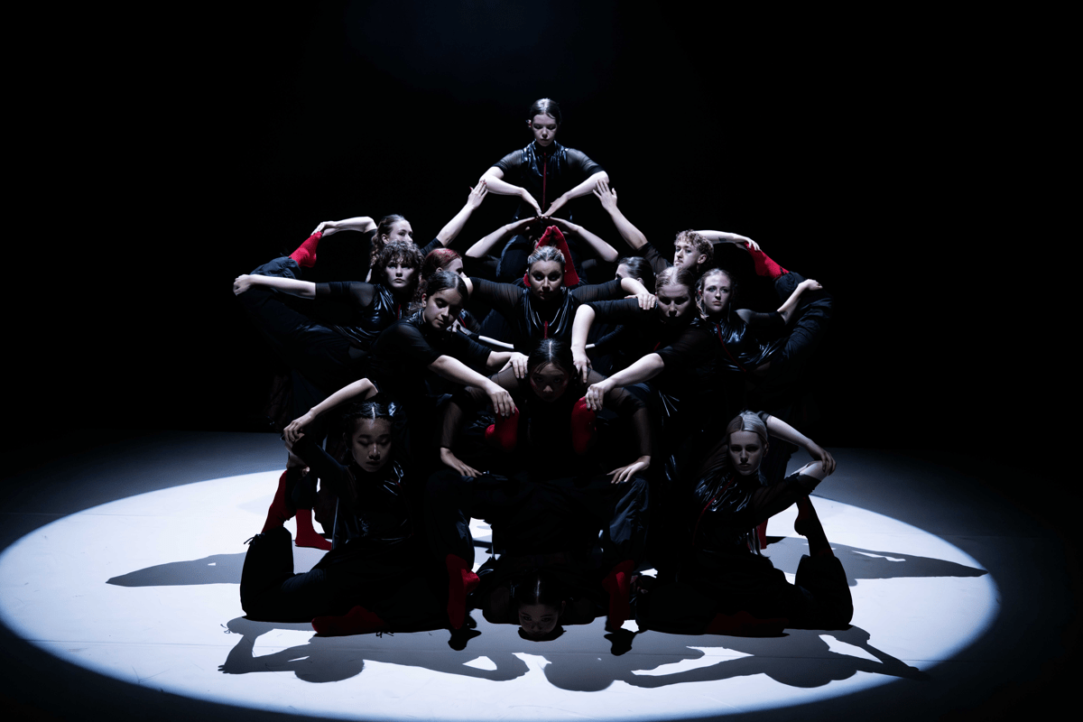 Image of a group of dancers huddled together on a stage wearing black clothing with red accents. They are looking forward, forming a loose geometric star-shape with their arms and legs. They are lit from above by a single bright white spotlight.