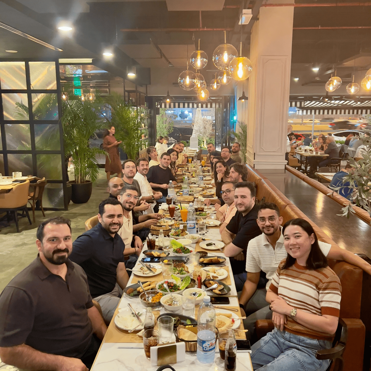 A large group of coworkers sit along a long restaurant table filled with dishes and drinks, smiling at the camera under warm hanging lights in a busy indoor setting.