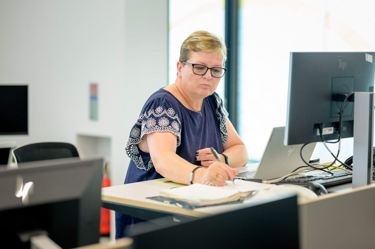 A woman working at a standing desk. There is a laptop in front of her and she's writing down something on a notepad.