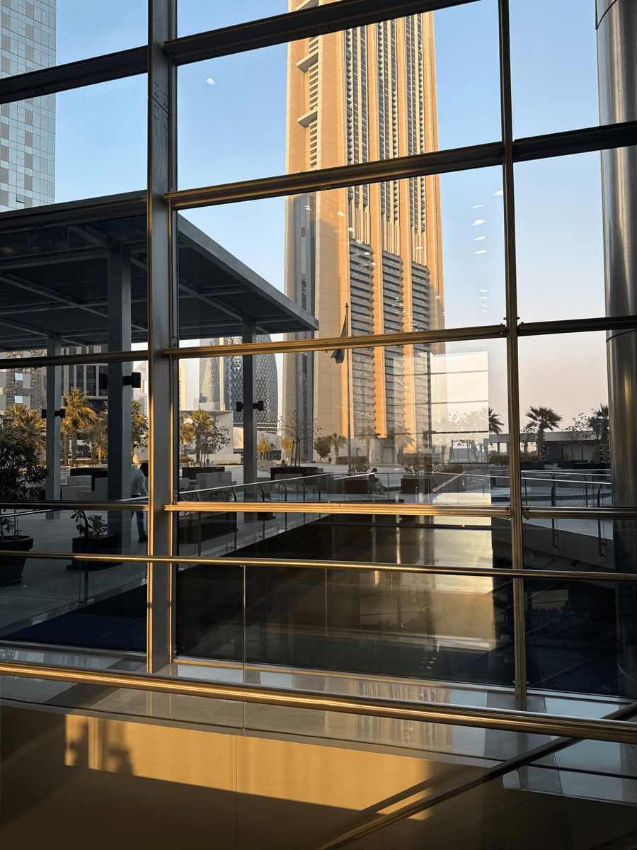 Golden-hour view through large glass windows toward a tall tower, with an outdoor terrace, palm trees, and reflections on the glass.