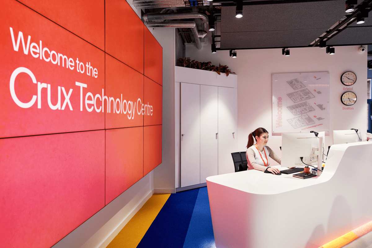 Receptionist at a white reception desk in a bright, modern lobby, with a large screen wall behind them showing the message ‘Welcome to Crux.