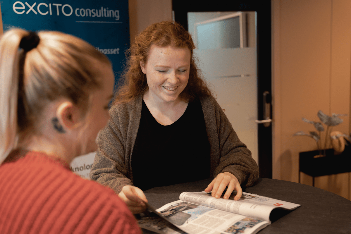 To kvinnelige smilende konsulenter fra Excito som står ved et bord og blar gjennom et fagblad. 