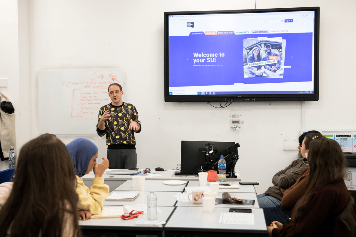 A presenter gestures enthusiastically while speaking to a group of seated students in a classroom setting, with a large screen displaying a "Welcome to your SU!" message and images of two individuals labeled as co-presidents.