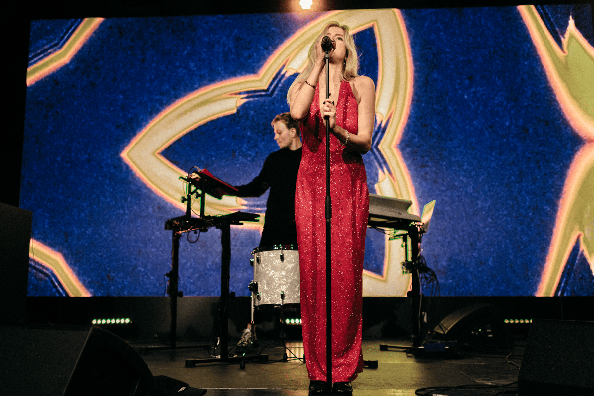  An artist singing on stage in a sparkly red dress with another person behind performing on electronic instruments 