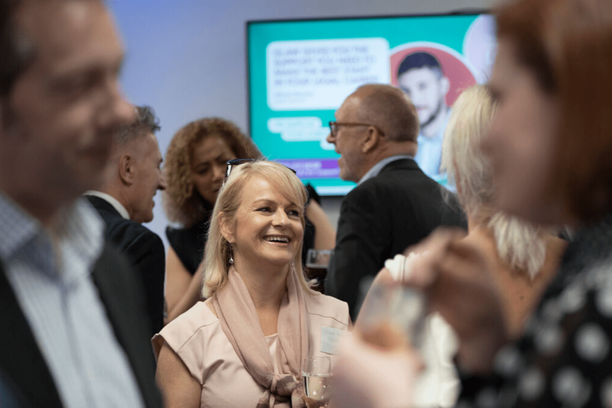 A woman with blonde hair smiles broadly while wearing a light pink top and a scarf at a social gathering, with a screen displaying text and an image in the background.