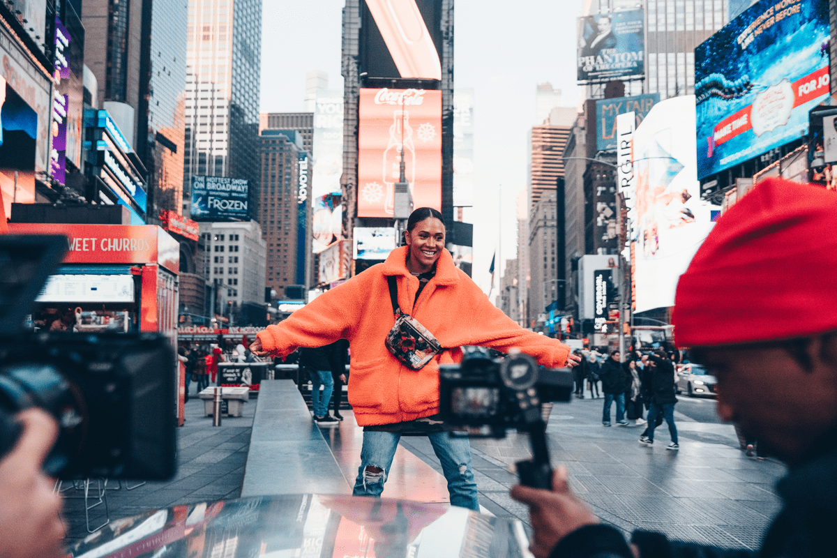 A creator in a bright orange jacket posing infront of the camera with her arms spread wide in the middle of a bustling Times Square in New York City