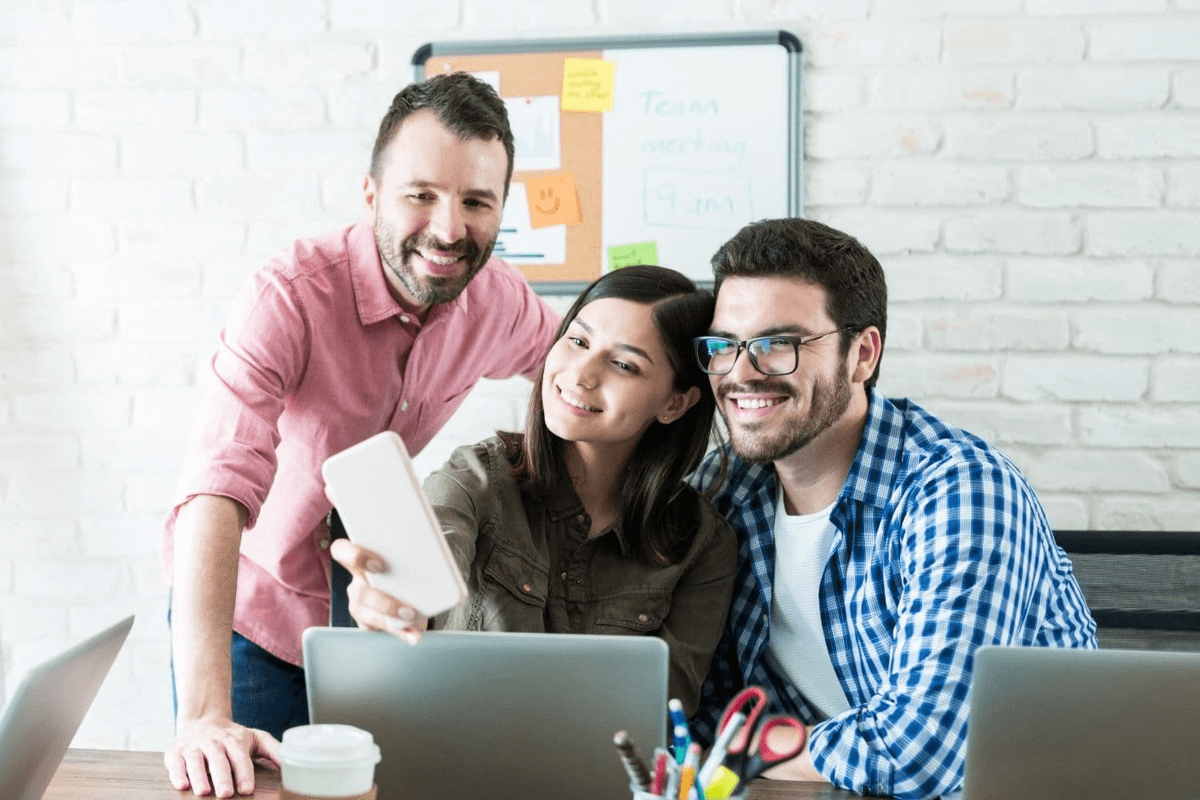 Team sitzt lächelnd vor dem Laptop und macht schaut auf ein Smartphone