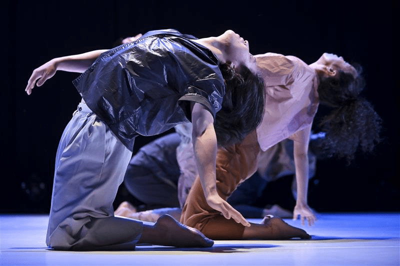 Image of two Korean dancers in profile wearing loose clothing kneeling on the floor of a stage, bending backwards, faces towards the ceiling.