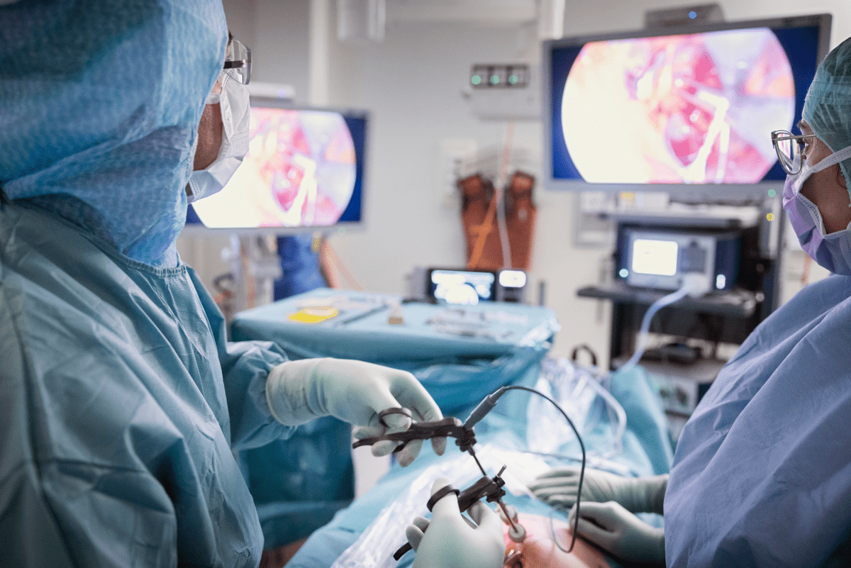 Två kirurger i skyddsutrustning utför en titthålsoperation i en operationssal med avancerad medicinsk utrustning och skärmar som visar bilder från ingreppet.