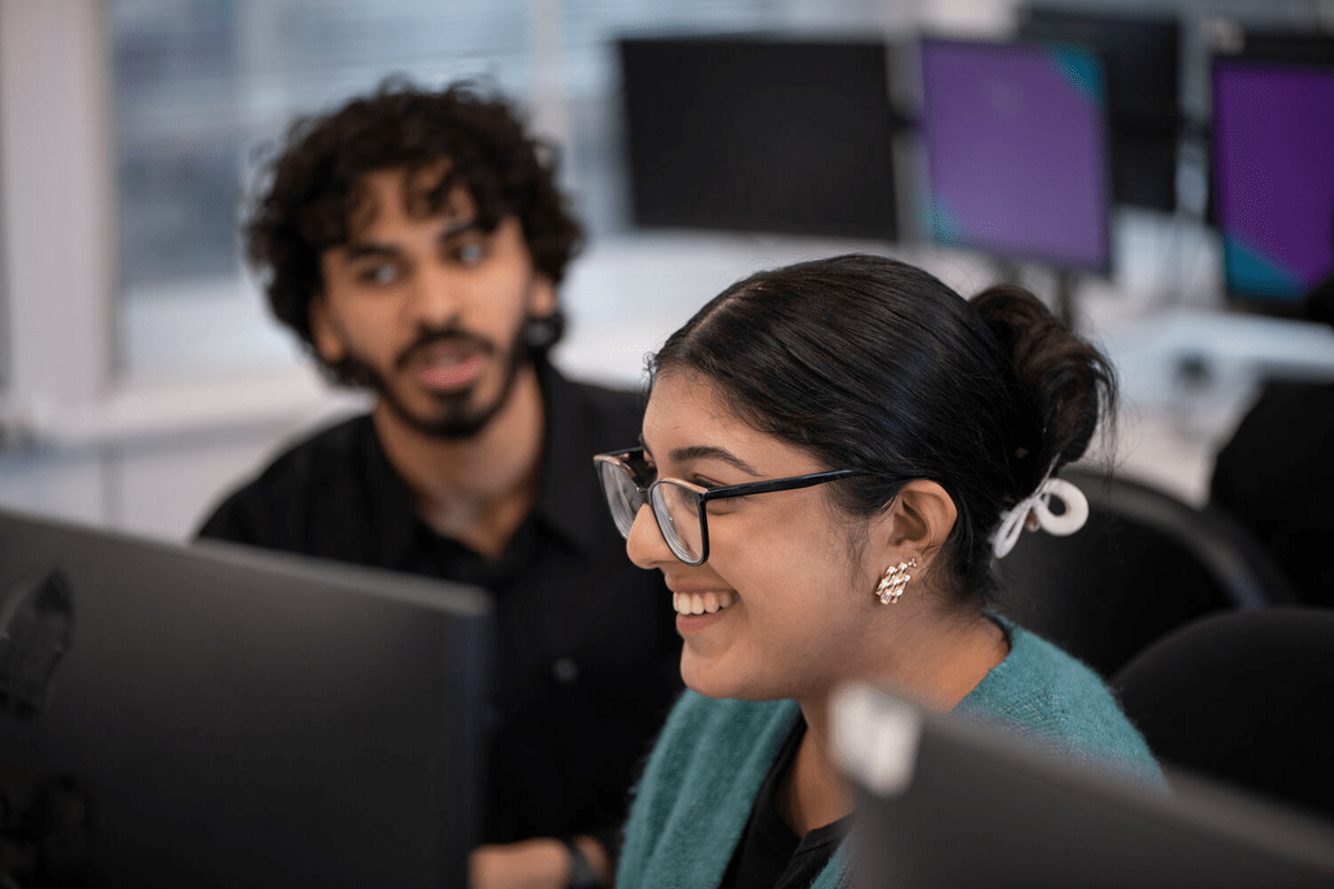 A woman in glasses smiles while looking at a computer screen, with a man in a black shirt in the blurred background.