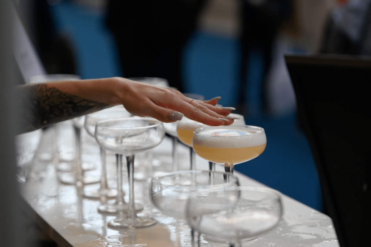 Martini glasses on a table. Some of them have a peach drink with a white head. A manicured female hand is spread over one of the glasses after dropping something on the top of the drink.