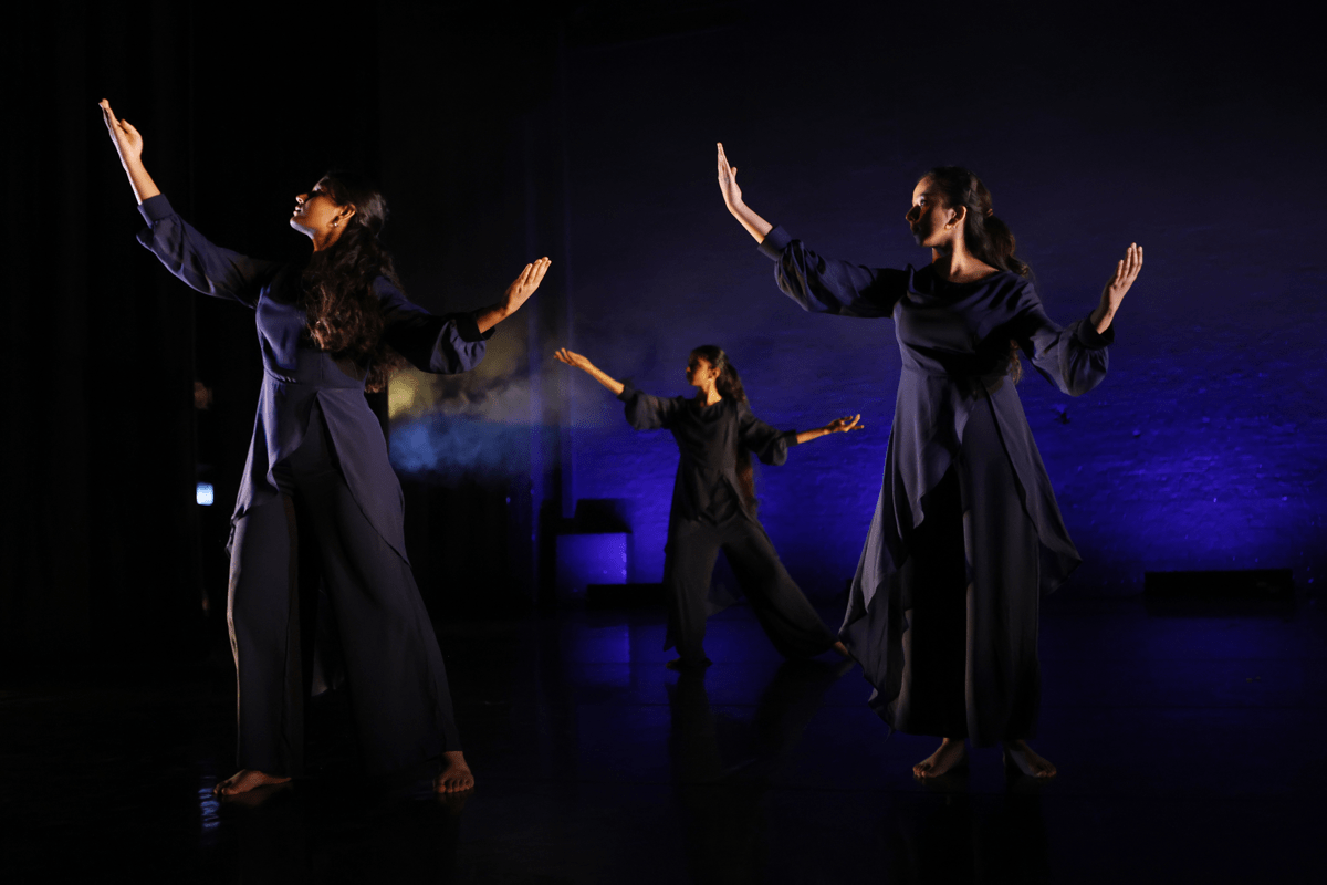 Image of three dancers on stage, backlit in blue lighting, wearing floor-length dark blue dresses, their arms are raised in the air with open palms.