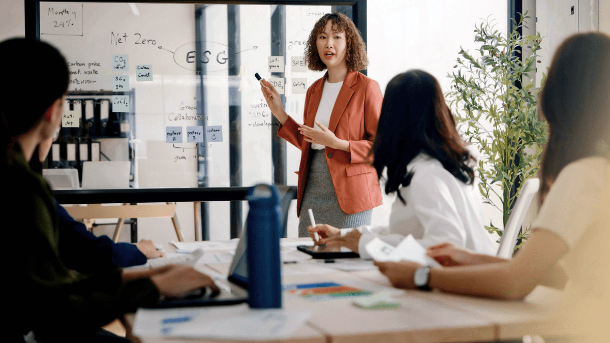 Three people sitting around a table listening to a woman talking beside a transparent glass whiteboard