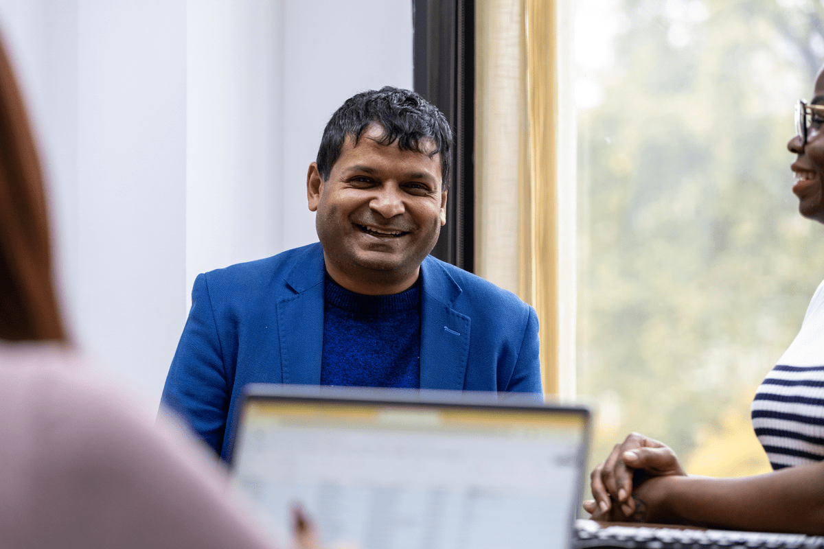 Man in a blue blazer smiling in conversation during a meeting near a window.