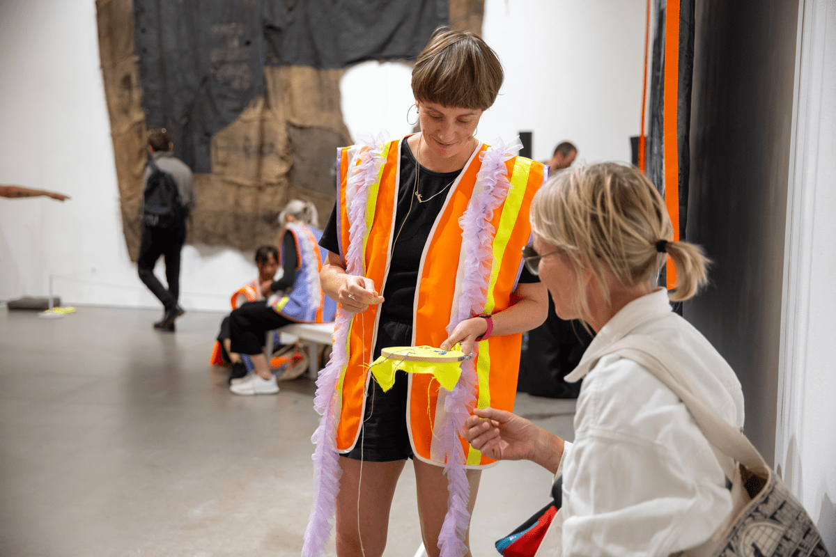 Image of a person in dark shorts and short sleeved top, wearing large hoop earrings, orange safety vest with yellow stripes, short brown hair, smiling, holding cross stitch out to a person with glasses, blonde hair tied in a ponytail, wearing a casual white collared shirt