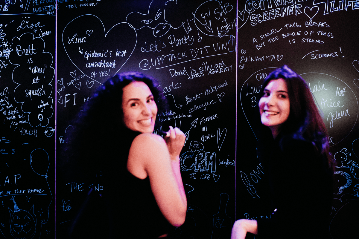 Two employees looking behind smiling at the camera whilst standing infront of a chalk board where employees have left messages for other employees to read