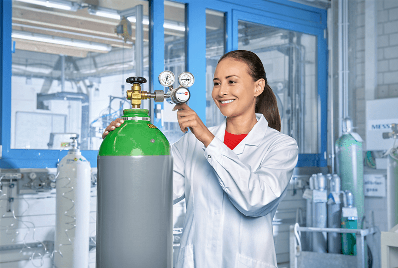 A woman wearing a lab coat stands with a gas cylinder, showcasing her role in a scientific research environment.