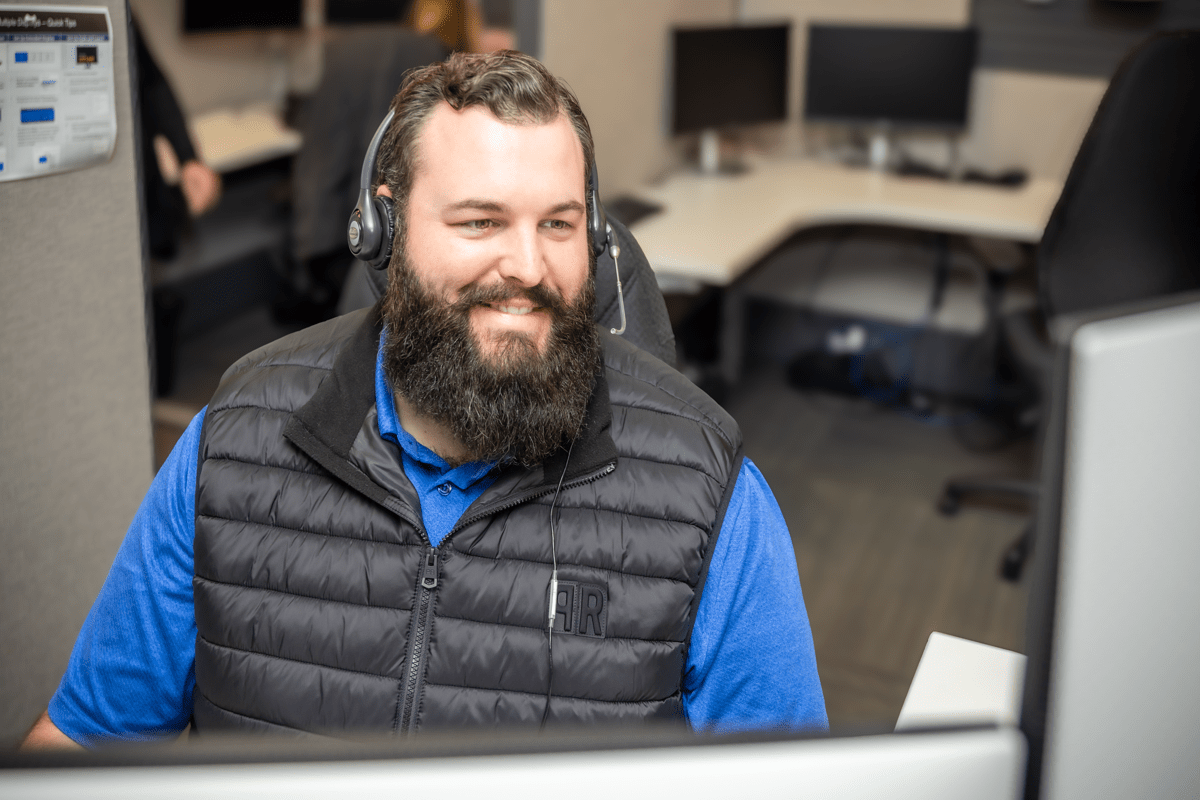 A smiling employee wearing a blue shirt and black vest works at a computer in a bright, modern office.