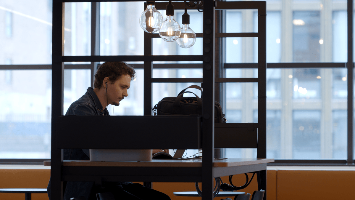 An employee wearing headphones and working on his laptop alone. Aesthetic, industrial-style light bulbs hang over the desk where they are sitting. In the background there are large windows letting lots of natural light in and showing the buildings outside