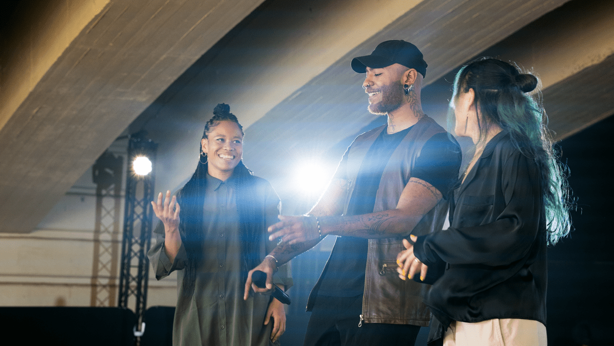 Three people stand together on a stage under an industrial-style ceiling. They appear to be performing or speaking to an audience. The person in the center, holding a microphone, smiles and gestures with both hands, while the two people on either side look at him and smile. Bright stage lights shine from behind them, creating a warm, energetic atmosphere.