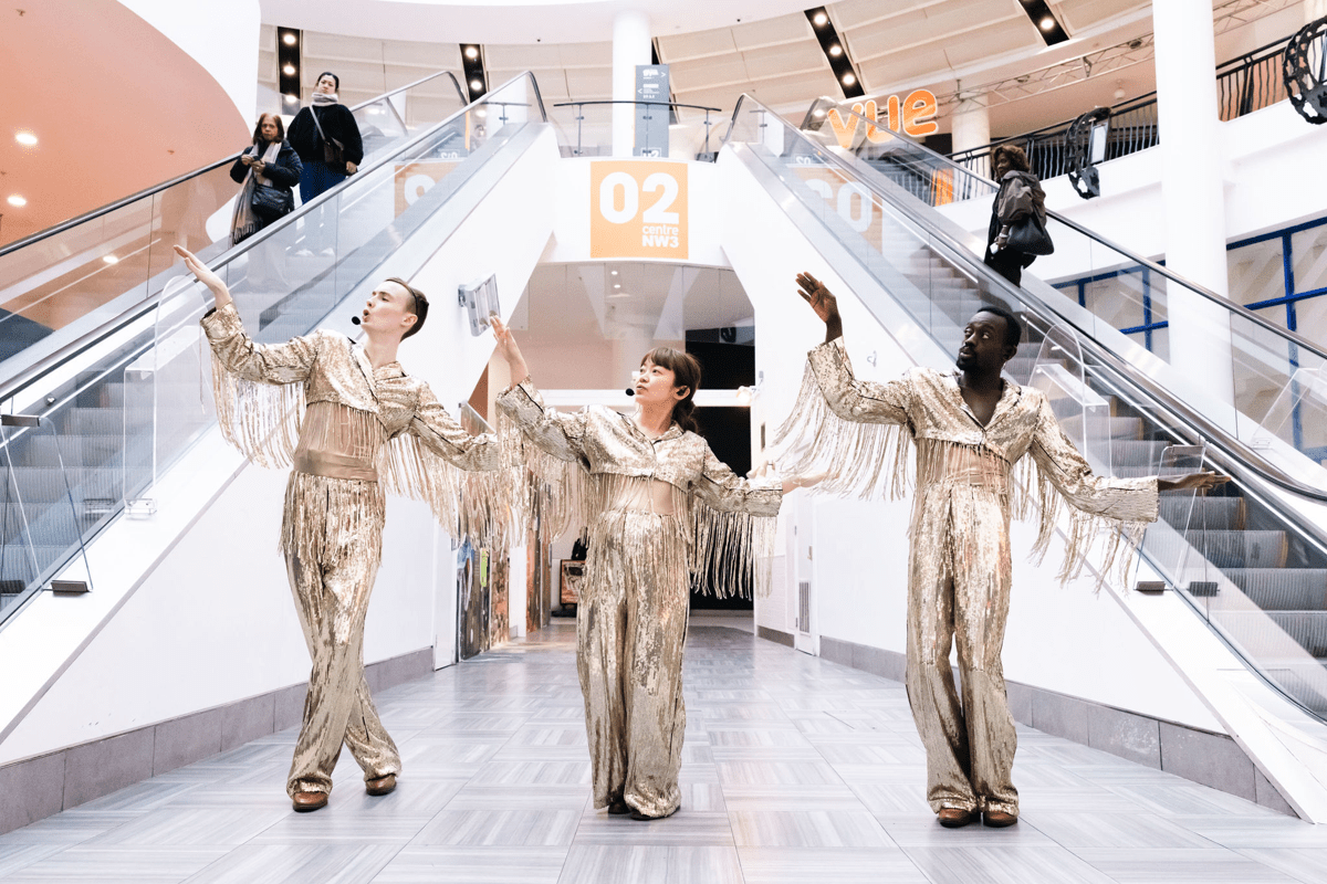 Three dancers in gold suits striking a pose in a shopping mall, between two escalators