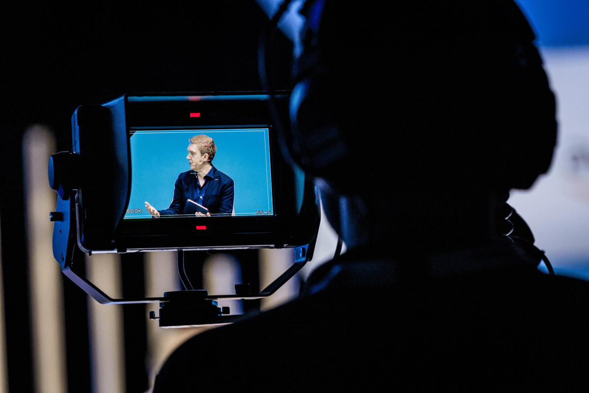 A backview of a cameraman who is in silhouette. On the camera screen you can see Nik Willetts in front of a light blue background. He is sitting and looking to his right.